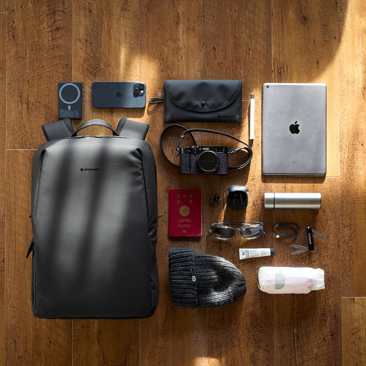 Artisan and Artist black travel backpack flat lay with camera, iPad, passport, power bank and daily essentials on wood surface, organised Japanese travel bag for Asia trips
